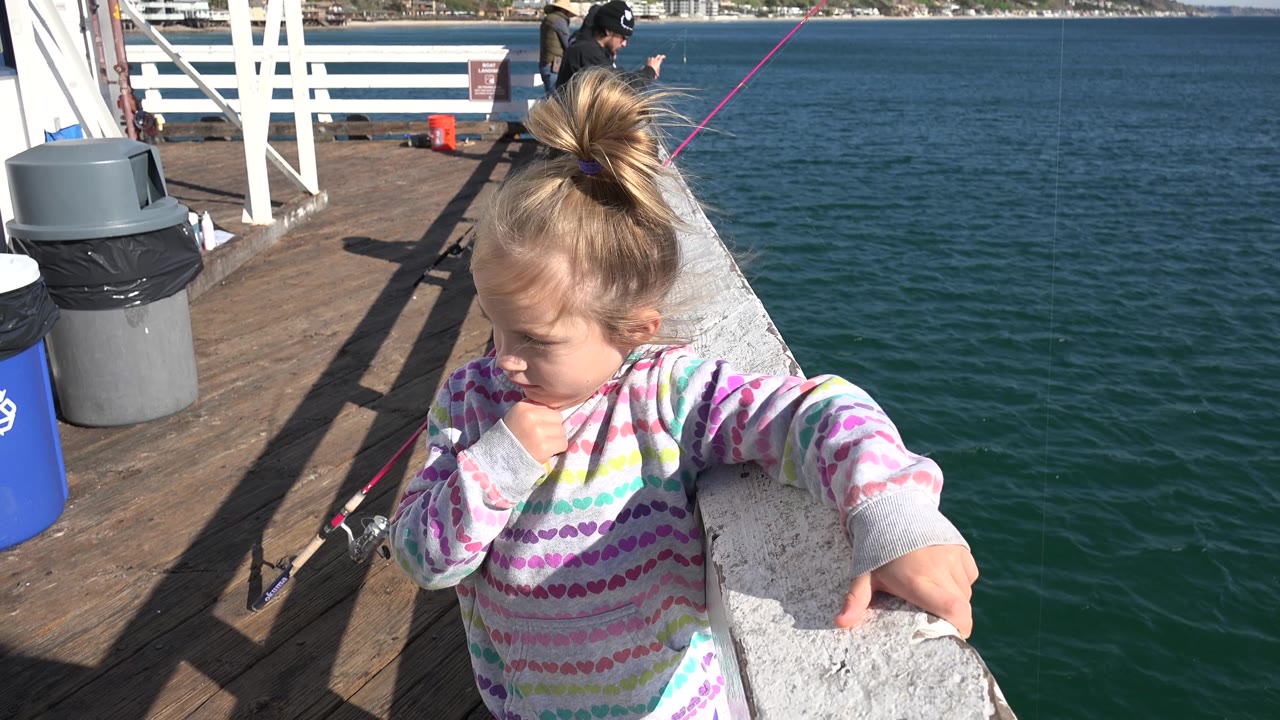 A young girl on the Malibu boat holds the rail while wind lifts her hair, with open water to the right and deck gear, bins, and fishing rods to the left.