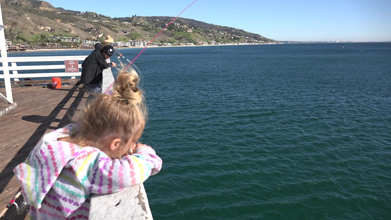 At 0:00 in Malibu, California, a young blonde girl in a colorful hoodie leans over the pier rail while adults fish beside blue ocean water and hillside coastline.