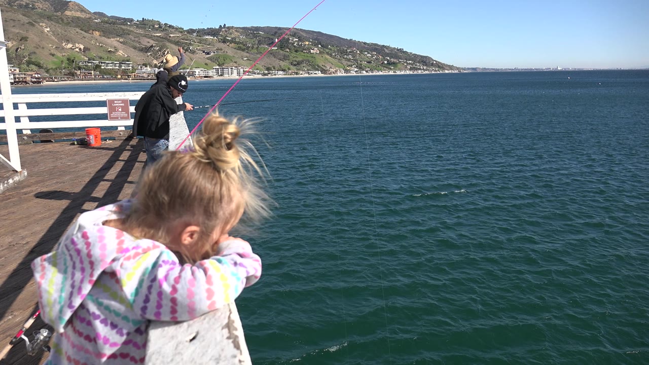 At 0:02 in Malibu, California, the girl looks downward from the left foreground while the adult anglers stand near the white railing and the ocean fills most of the frame.