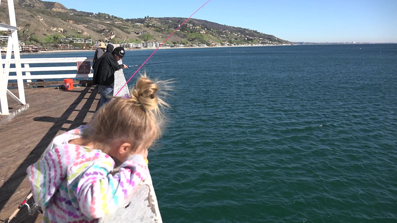 A young girl in a white striped jacket leans near a white pier railing in Malibu, with blue ocean, hillside coastline, and fishermen in the background.