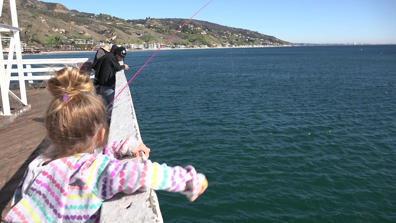 Seen from behind, the girl holds the white railing on a Malibu pier while the ocean stretches across the frame and the pink fishing line angles upward.