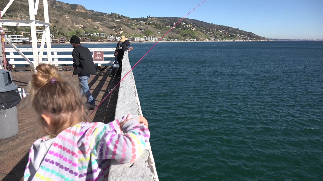 Child beside a pier railing with ocean and hills beyond.
