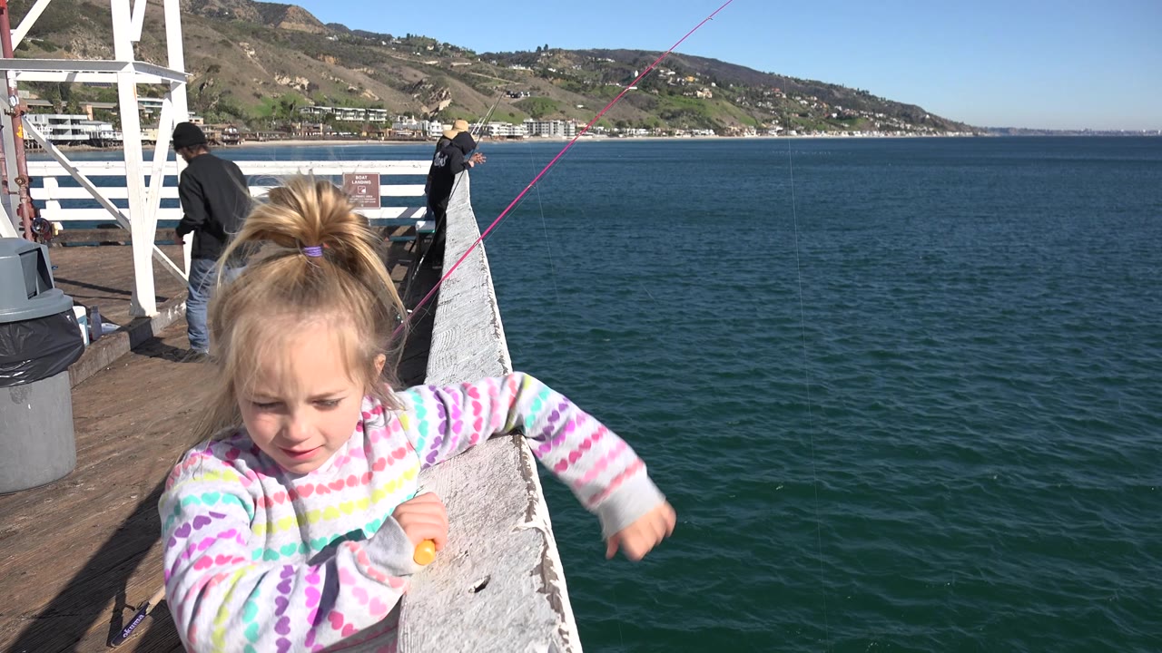 A young girl faces the camera beside a white Malibu pier railing, with bright sun on her striped hoodie and open blue water behind her.