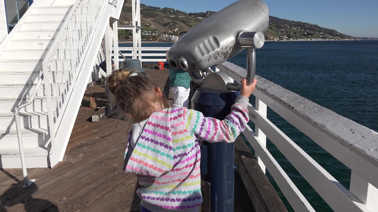 The girl stands close to the grey viewing telescope on a Malibu pier, with her hand on the side handle and the ocean horizon visible beyond the railing.