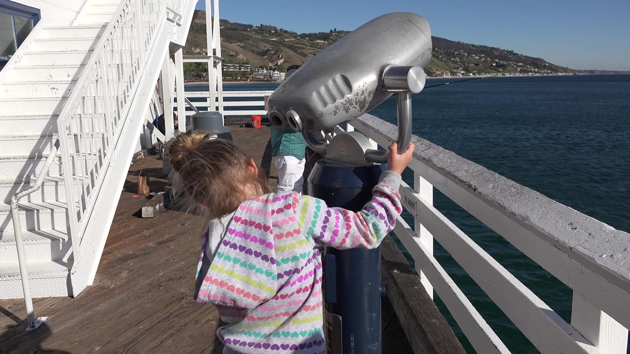 A young girl remains beside the mounted grey telescope on the Malibu pier, framed by white railings, dark blue water, and a hillside shoreline in bright daylight.