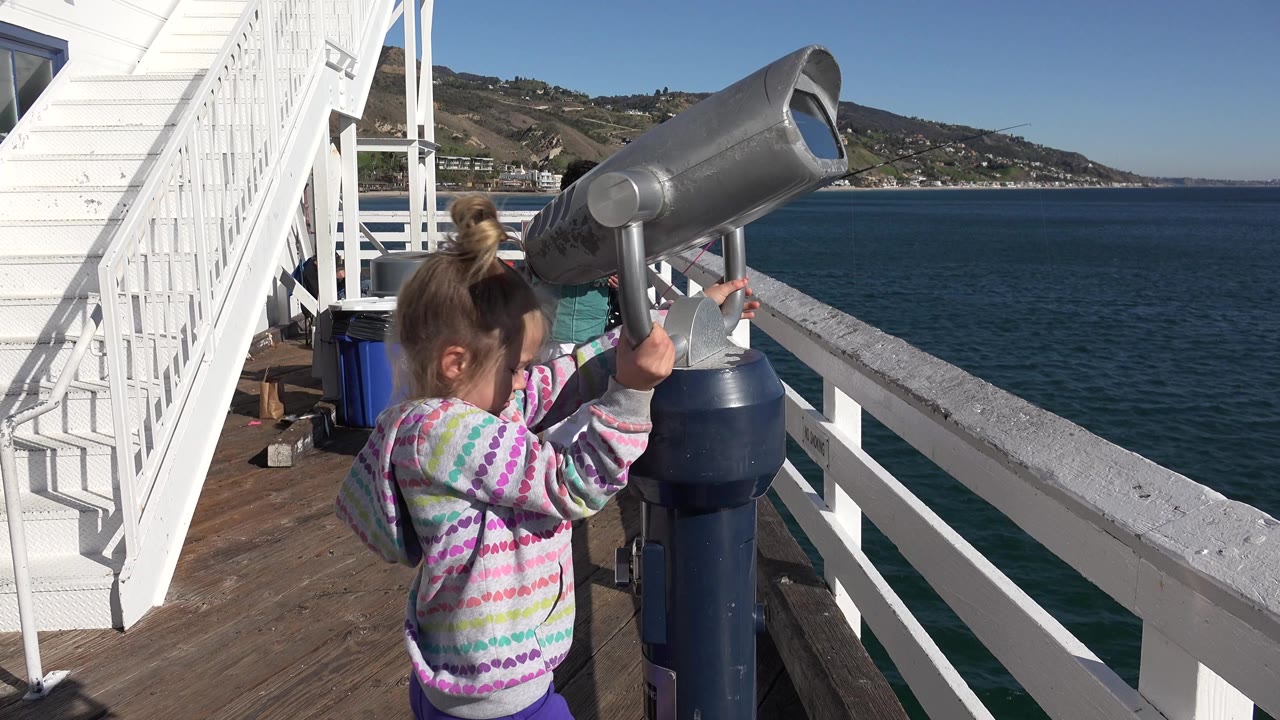 At the start of the Malibu clip, a young girl stands close to a metal pier viewer with blue ocean and a hillside shoreline beyond the railing.