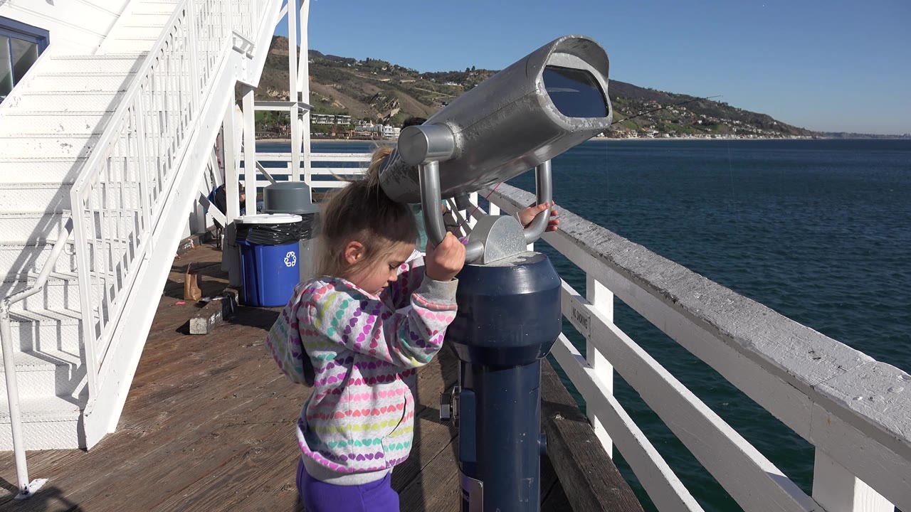 The girl lowers her face near the side of the viewer on the Malibu pier, with sunlit water, railings, and the sloped shoreline in the background.