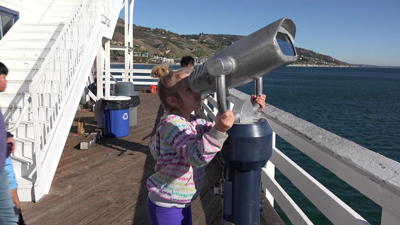 In Malibu, California, a blond girl looks into a large metal viewing telescope on a ferry deck while white stairs, railings, blue water, and distant hills fill the background.