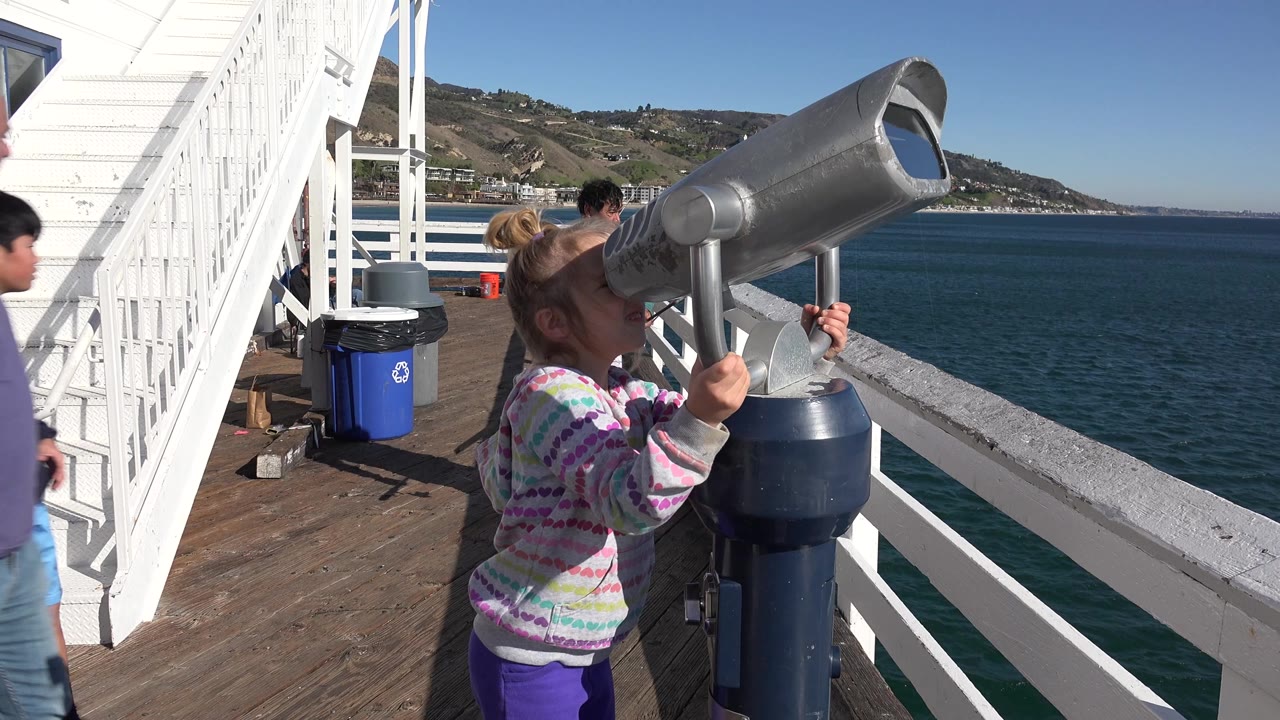 The Malibu ferry deck view holds on the girl at the telescope, with adults partly visible nearby, white ferry stairs to the left, and open blue water behind the railing.