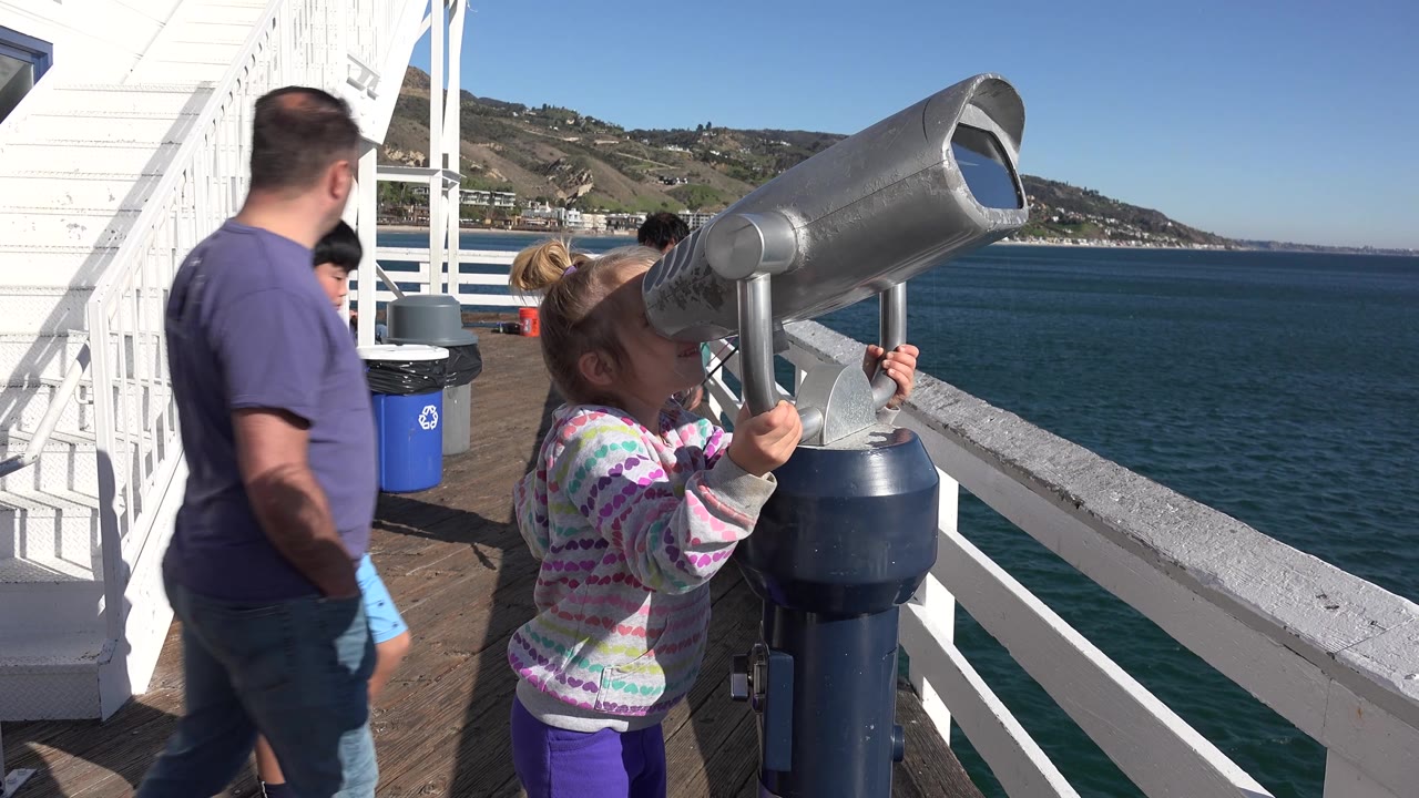 A man in a purple shirt passes between the camera and railing while the blond girl continues using the telescope on the Malibu ferry deck with ocean and hills beyond.
