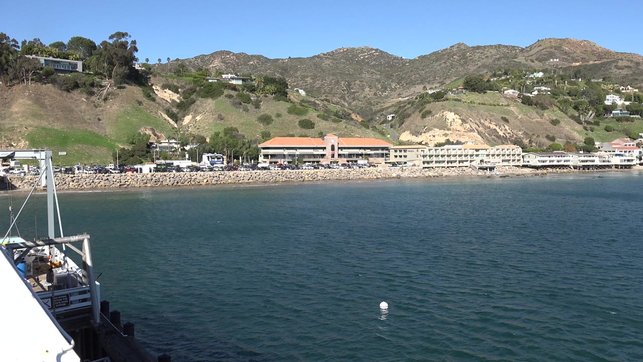 Malibu ocean water fills the foreground with beachfront buildings and green hills beyond, a boat or pier edge at left, and no visible people.