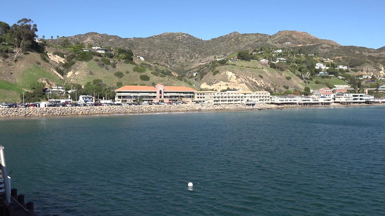 Opening Malibu frame with blue water in the foreground, low resort buildings along the shore, hillside homes in the background, and no visible people.