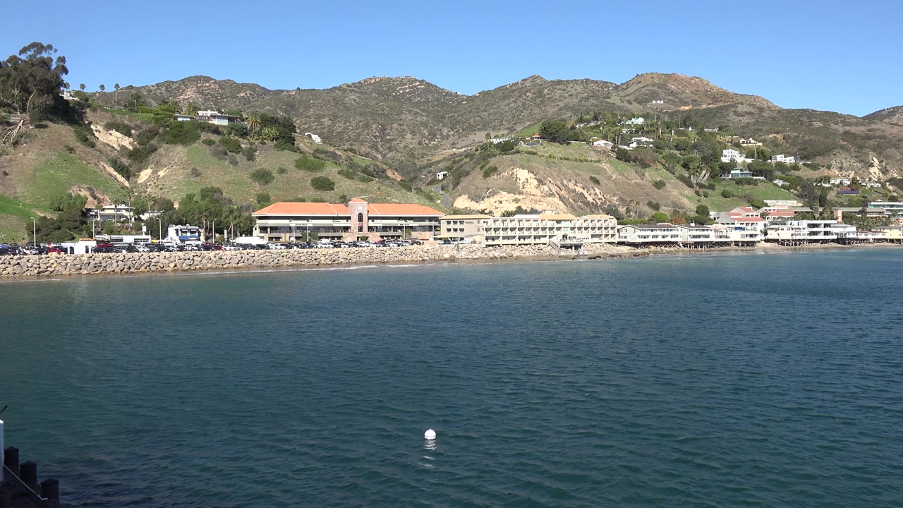 Two-second Malibu frame showing more open water to the right, the resort line across the shore, layered hills behind it, and no visible people.