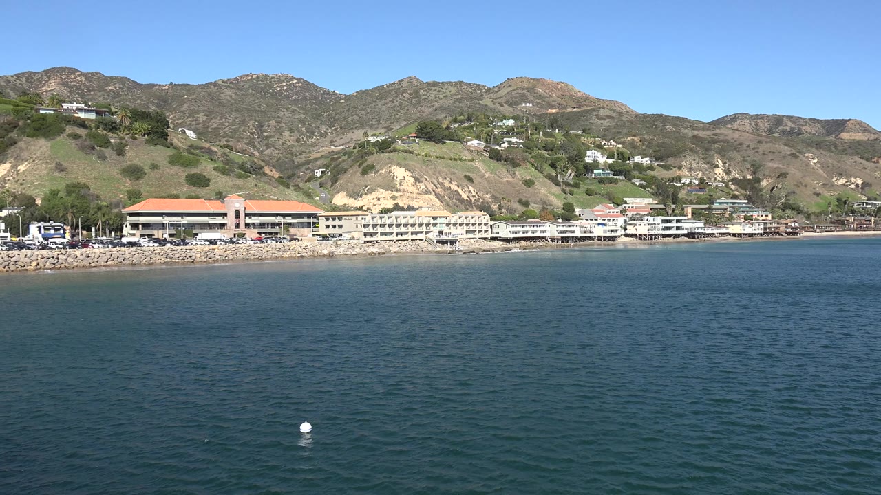 Wide Malibu, California view across rippling blue water toward low shoreline buildings with red tile roofs, rugged hills, scattered hillside homes, and no visible people.