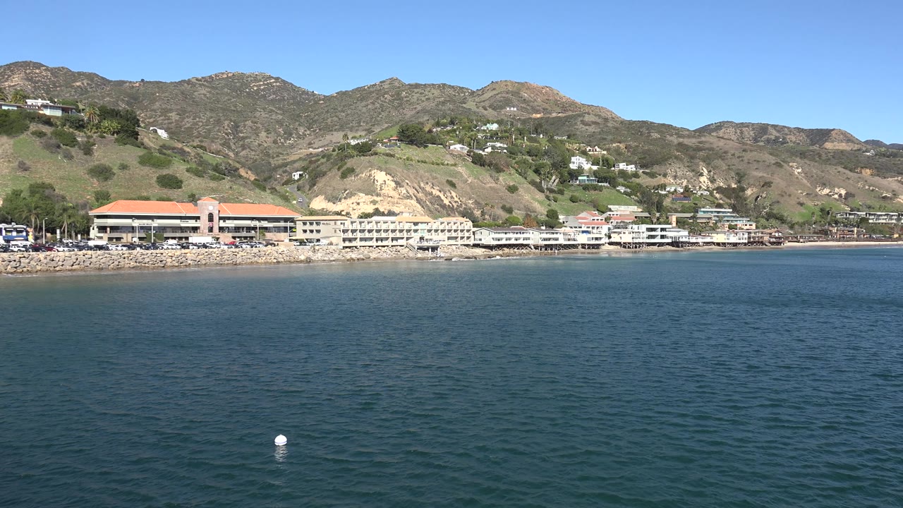 Wide Malibu shoreline frame with dark blue water in the foreground, a buoy near the lower center, oceanfront buildings, pale cliff cuts, hillside residences, and no visible people.