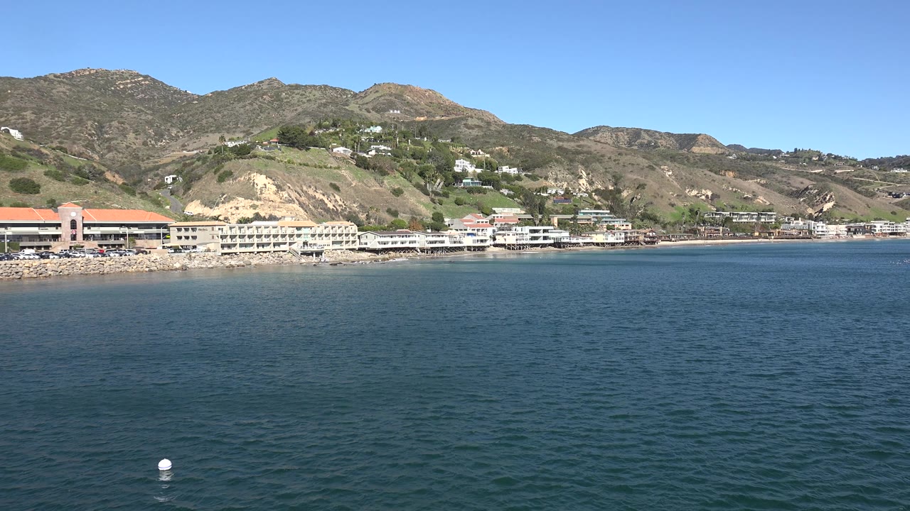 Sunny Malibu coast is seen over textured blue water, with a small buoy near the lower edge, low buildings on the beach, layered hills inland, and no visible people.