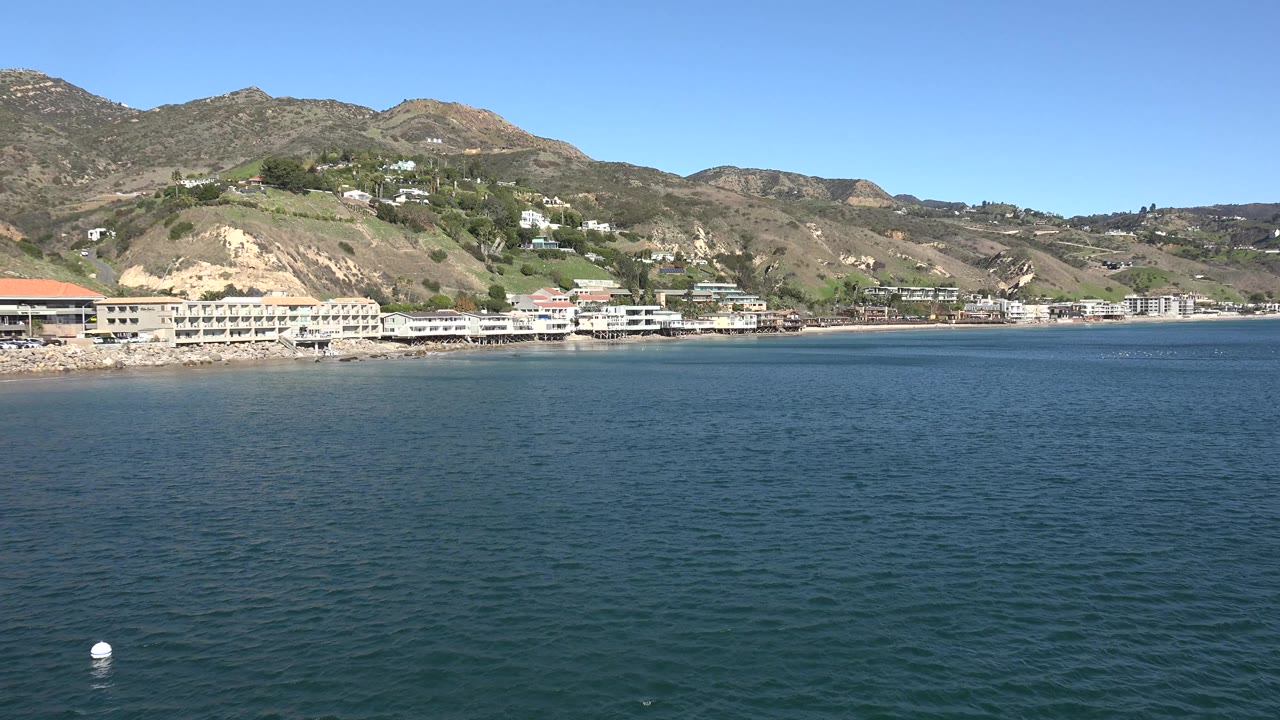 Wide view across blue-green water toward Malibu shoreline buildings and rolling hills under a clear blue sky, with no visible people.