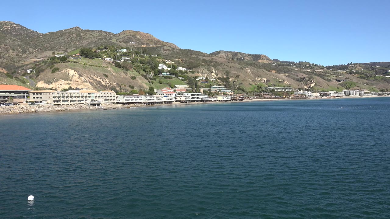 Ocean foreground with a white buoy, shoreline buildings, and hillside homes.