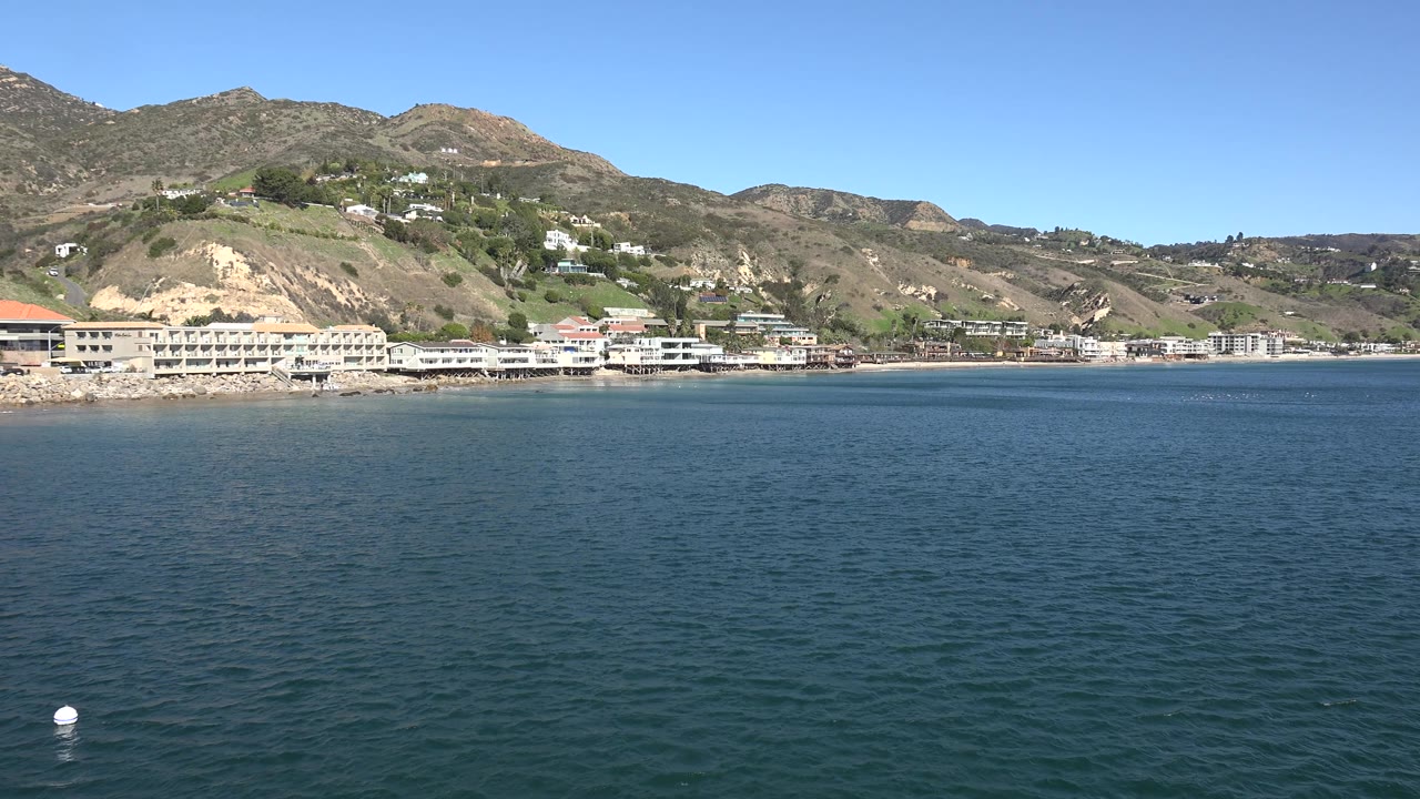 Calm water stretches toward Malibu beachfront structures and dry rolling hills, with a clear blue sky above and no visible people.