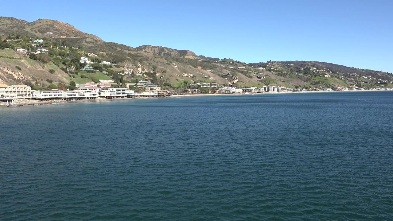 Blue water in foreground with coastal homes below hills.