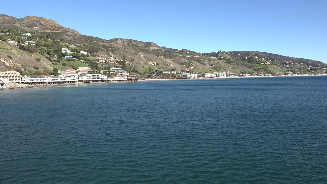 A broad water-level view shows rippled ocean leading to a Malibu hillside neighborhood and low shoreline buildings, with no visible people.