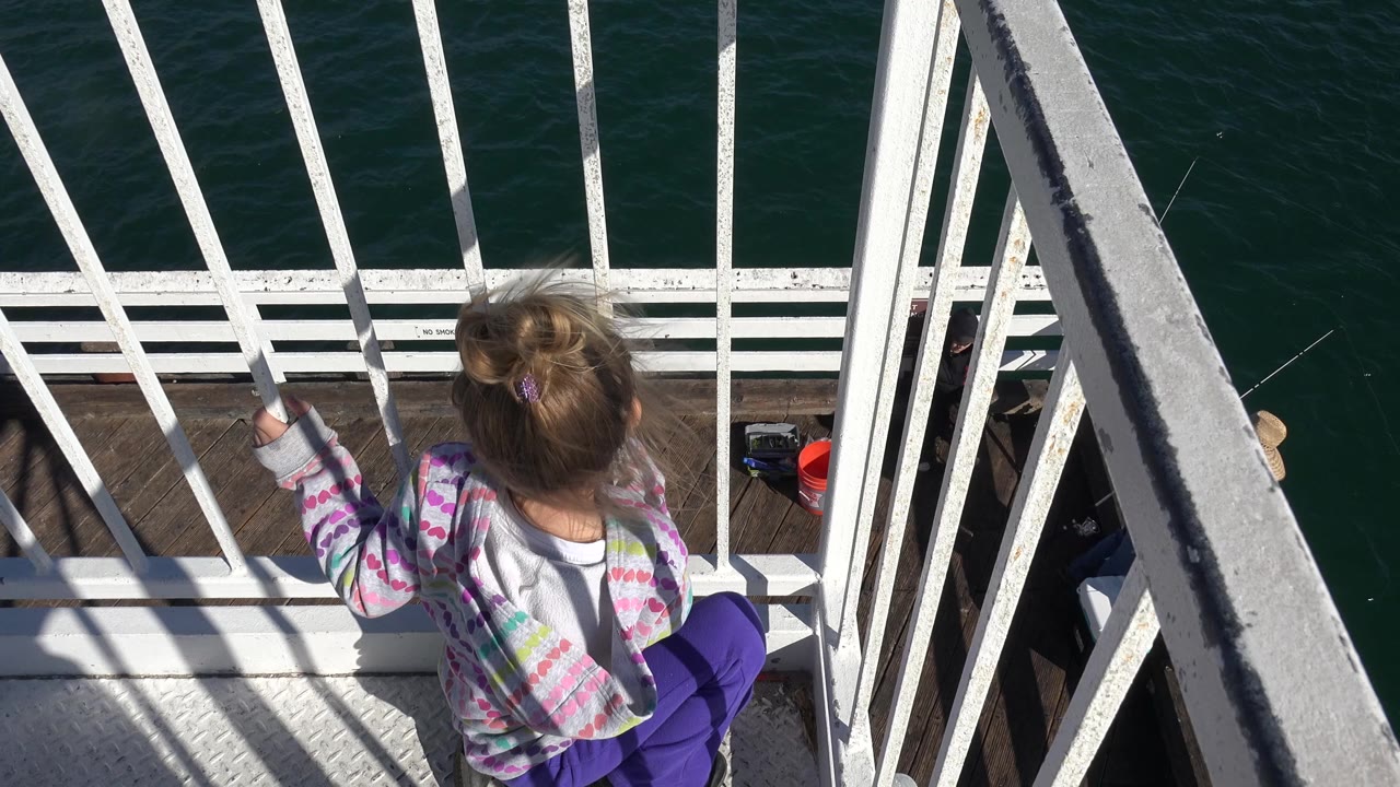 The Malibu balcony view holds on the girl from behind as sunlight strikes the white railings and the ocean fills the background.