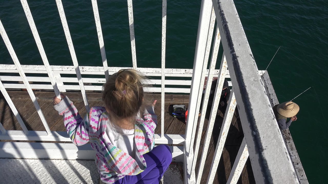 At 0:00 in Malibu, California, a young girl seen from behind sits near white balcony rails above dark green ocean water, with a lower deck partly visible at right.
