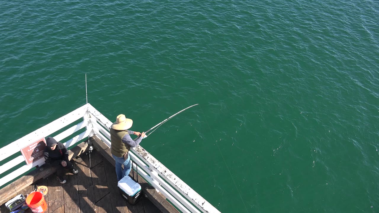 At 0:00 in Malibu, a man in a wide hat stands at the corner of a white-railed deck with a fishing rod while another person sits nearby above green ocean water.