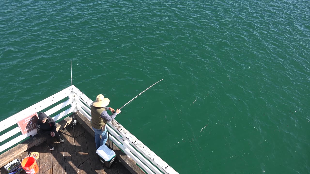 At 0:02 in Malibu, two people remain on the deck corner, with the fisherman facing the water and the seated person beside the rail as sun catches ripples below.