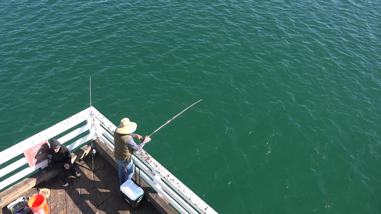 Deck corner with seated person and fisherman beside green water.