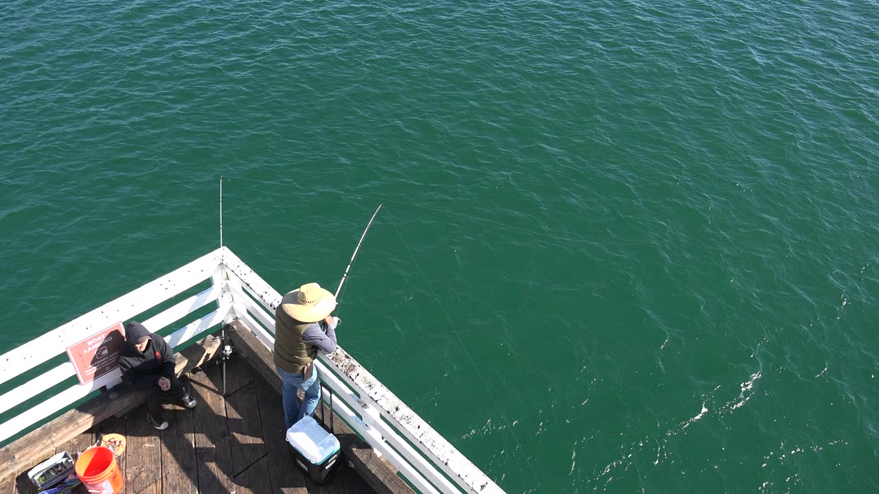 At 0:00 in Malibu, California, a person in a straw hat stands by a white pier railing with a fishing rod while another person sits nearby above green ocean water.