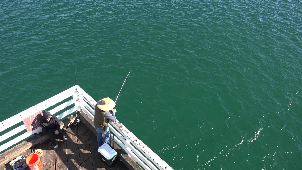 At 0:01, the straw-hatted fisher remains at the white railing in Malibu as small bright ripples appear in the green water beside the pier corner.