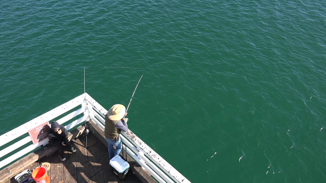 The Malibu pier corner remains in view as the standing angler in a straw hat faces the water and the seated person stays near the railing.