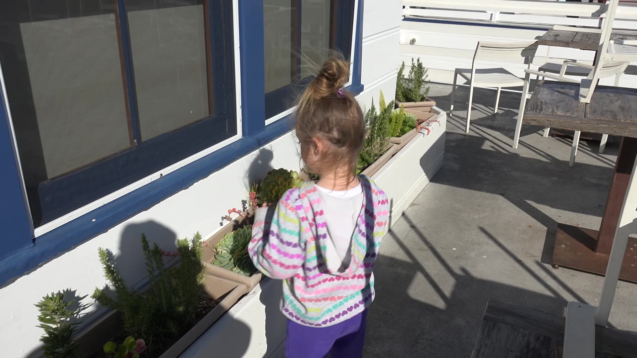 In Malibu, California, the girl turns more toward the planter, showing a partial side profile while white chairs and a dark wooden table sit behind her on the patio.