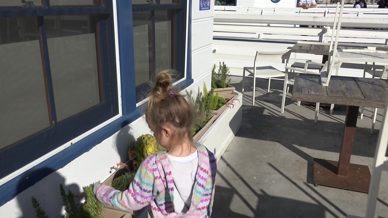 At 0:00 in Malibu, California, a young child seen from behind reaches toward planter greenery beside blue-trimmed windows, with white railings and a patio table in the sunlit background.