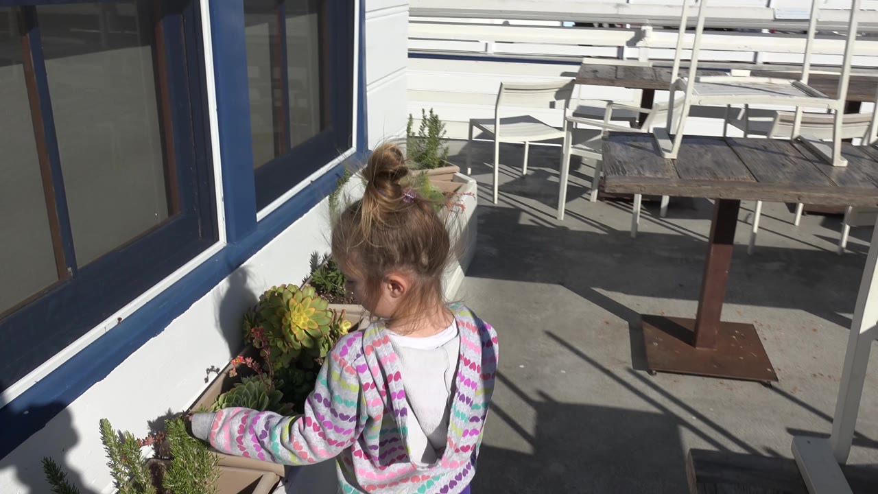 At 0:02 in Malibu, California, the child faces the planter from the side, with larger succulent leaves near her hand and sunlit railings, chairs, and a dark table behind her.