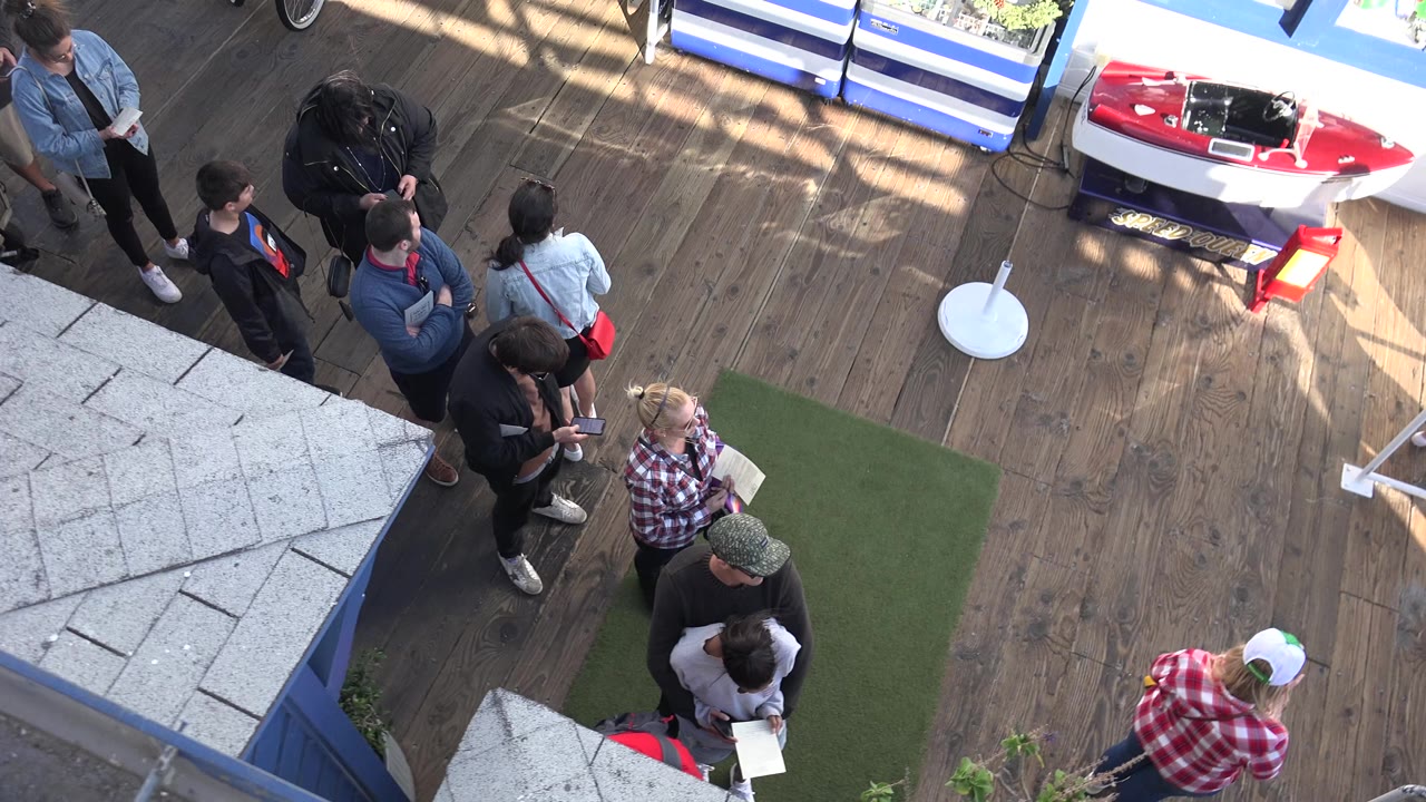 High-angle Malibu frame showing several adults standing on a wooden deck near artificial turf, a red kayak, and striped blue-and-white structures.