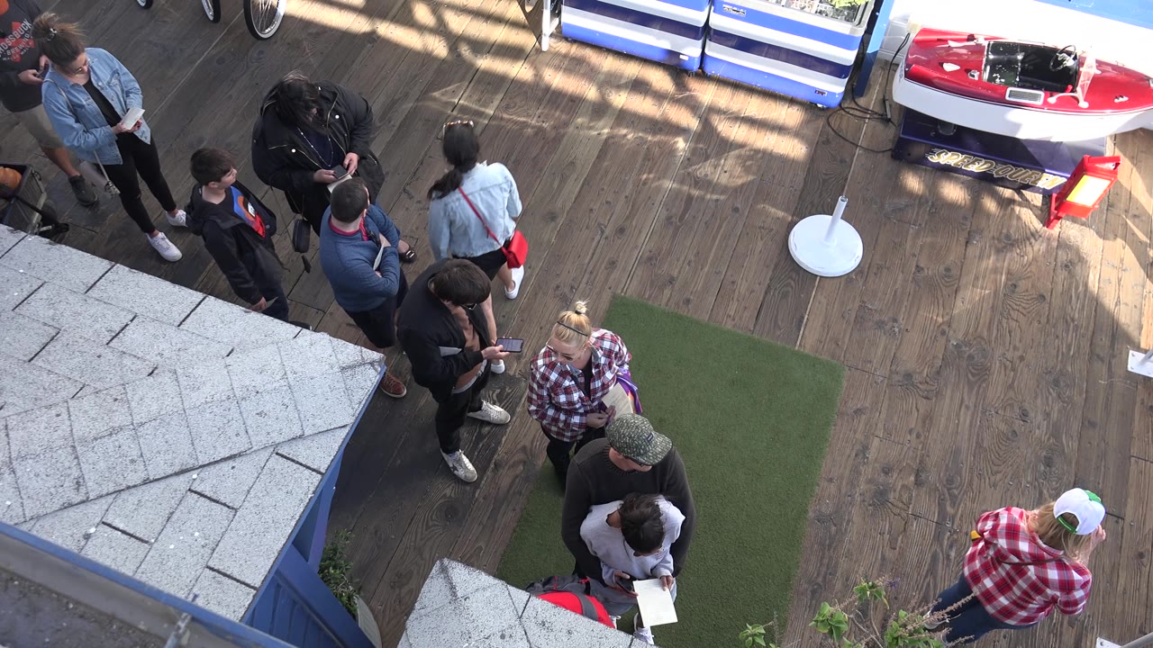 High-angle Malibu frame with adults moving slightly around the deck, a woman in a plaid shirt lower right, and the red kayak partly visible above.