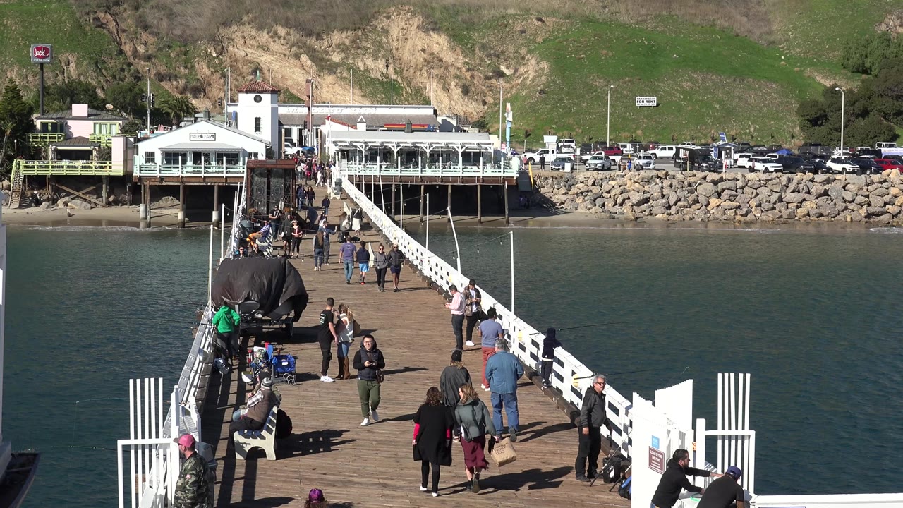 At 0:00 in Malibu, people walk along a wooden bridge with white railings over green water, with a waterfront building, parked cars, rocks, and grassy hillside beyond.