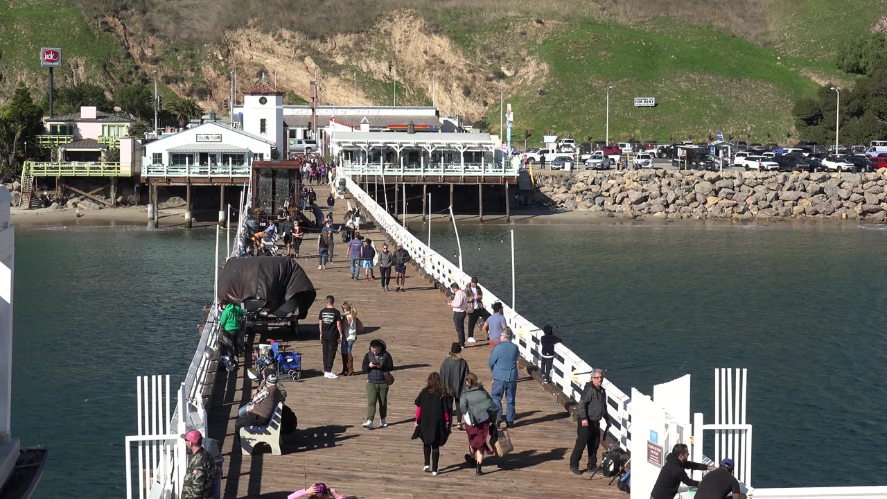 At 0:02 in Malibu, several people continue across the wooden bridge while the white railing angles toward waterfront buildings, parked cars, rock shoreline, and a grassy hillside.