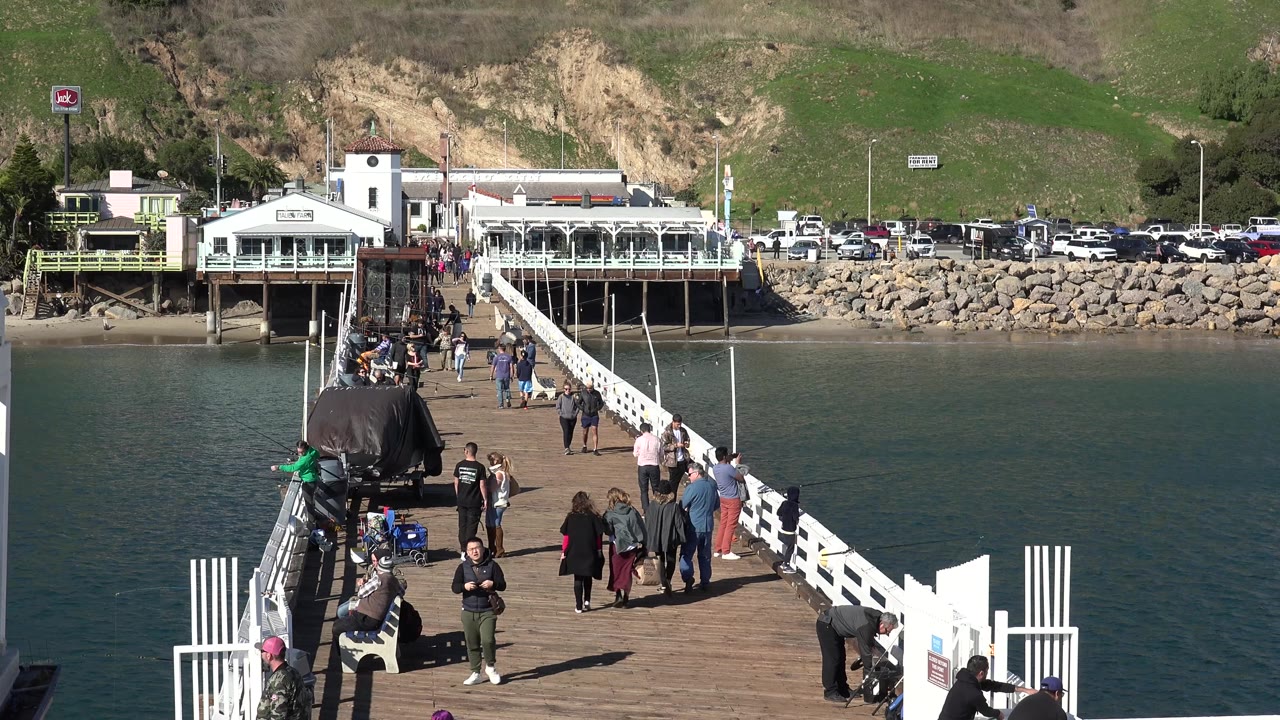 People move along a wooden pier in Malibu, California, with white railings, teal water, shoreline buildings, parked cars, rocks, and a sunlit green hillside in the background.