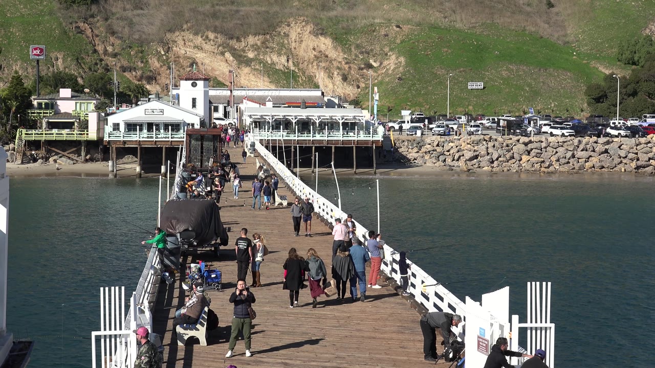 People walk and pause on the Malibu pier as bright sun hits the white railings, with teal water flanking the walkway and shoreline structures, cars, rocks, and hillside behind them.