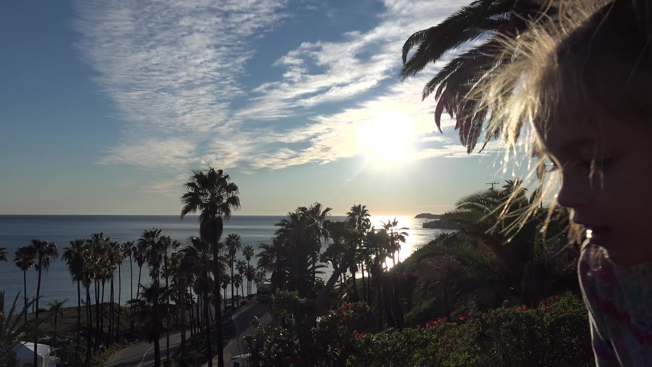 Child's backlit face in foreground with ocean and palms behind.