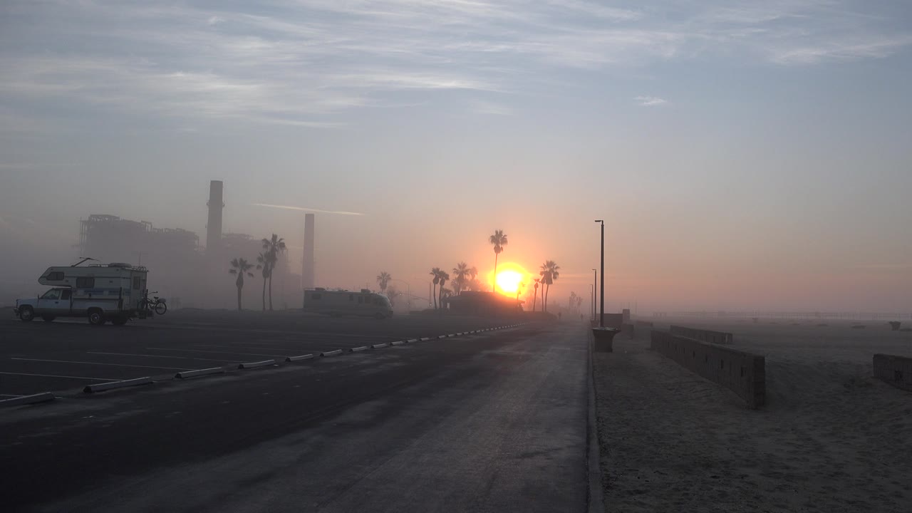 A Huntington Beach sunset sits just above the horizon, with palm trees clustered near the sun, an RV on the left side of the road, and no visible people.