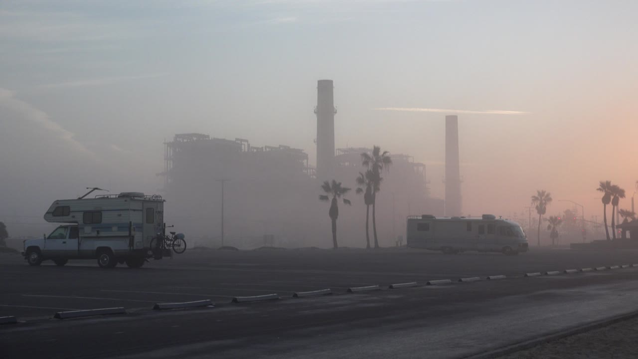 Huntington Beach, California frame with a camper vehicle at left, another RV at mid-right, palm trees, hazy industrial towers, peach horizon light, and no visible people.