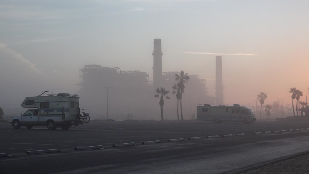 Huntington Beach, California frame holding on the same paved foreground, left camper, center-right RV, palms, industrial structures, gray haze, and no visible people.