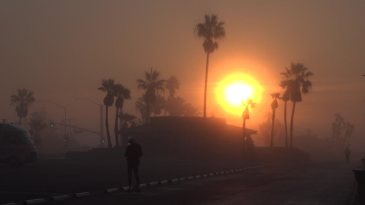 Opening frame of a silhouetted man on an empty road in Huntington Beach, California, with palm trees and a bright orange sun behind a dark low building.