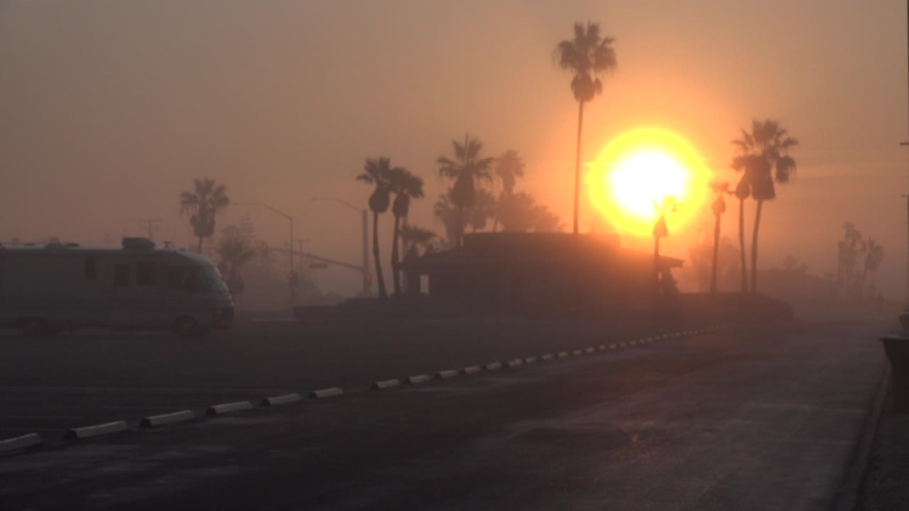 At 0:00 in Huntington Beach, California, the low orange sun glows through haze behind palm silhouettes, with a dark curb and a partial vehicle at left and no visible people.