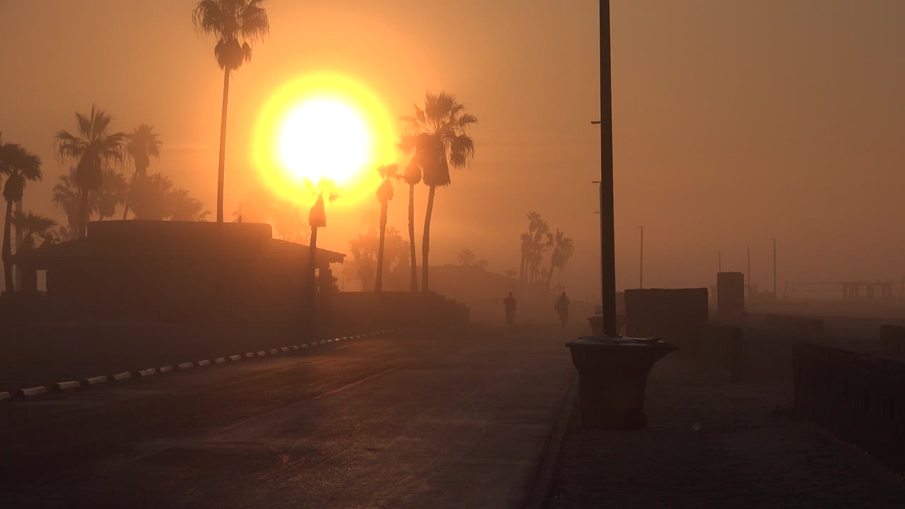 At 0:00 in Huntington Beach, a large orange sun sits behind silhouetted palms and a dark boardwalk-side structure, with an empty paved path and no visible people.