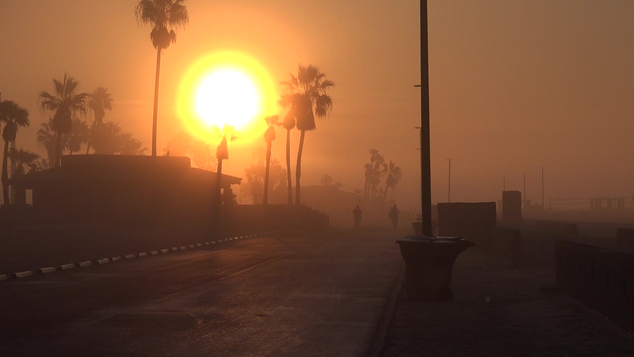 At 0:01 in Huntington Beach, the sun remains high in the orange haze behind clustered palms, with the boardwalk path and foreground pole framing an empty scene with no visible people.
